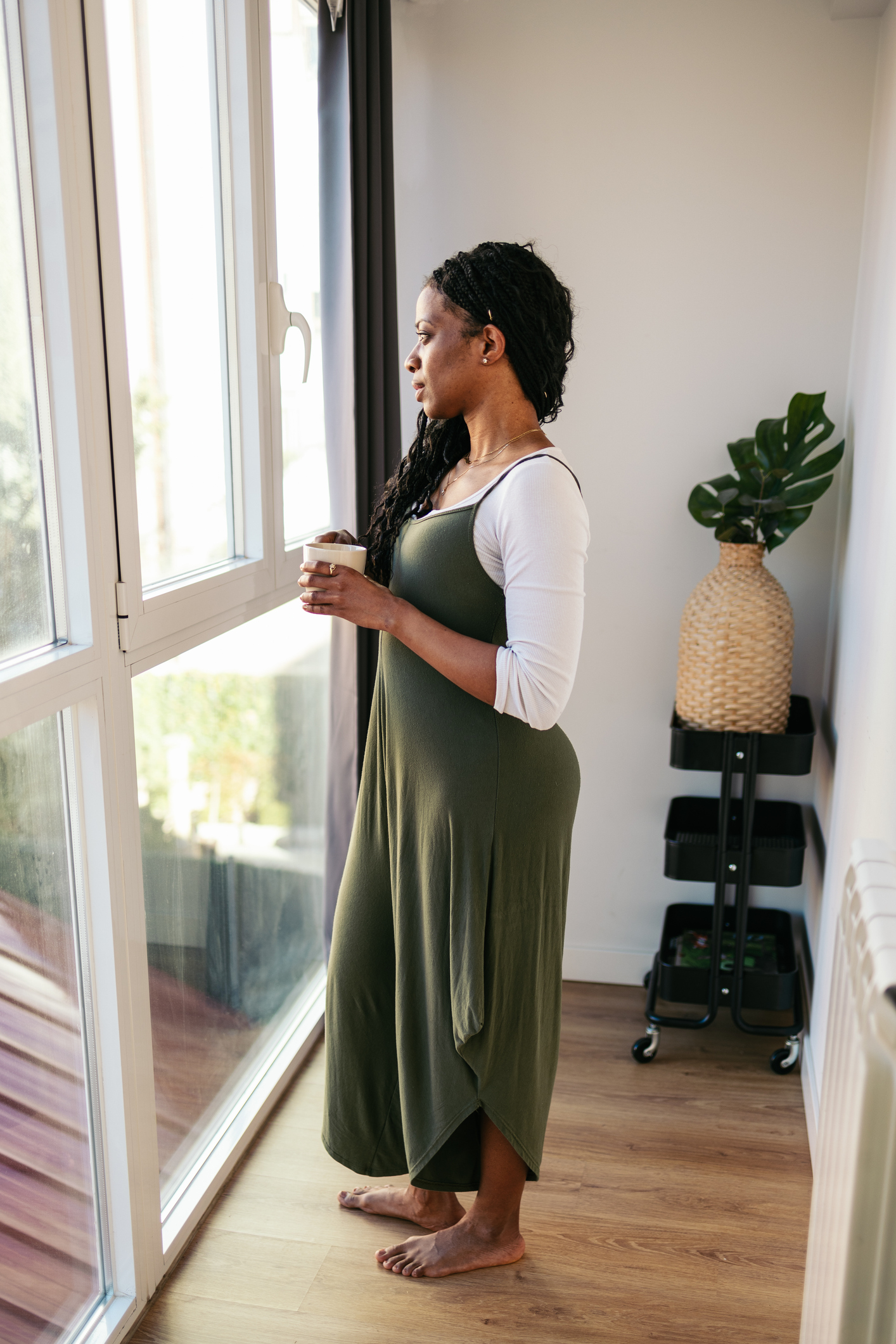Young woman holding a cup of coffee while looking out the window in her modern apartment