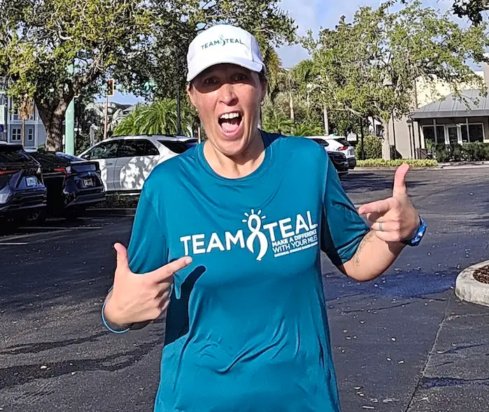 Ovarian cancer survivor Hollie Nelsen in her Team Teal shirt and cap pointing to the Team Teal logo during a daytime training run for the 2026 Boston Marathon