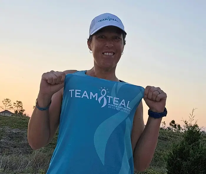 Hollie Nelson holds up her Team Teal racing shirt with both fists raised in excitement at sunset, days before running the Boston Marathon for the National Ovarian Cancer Coalition.
