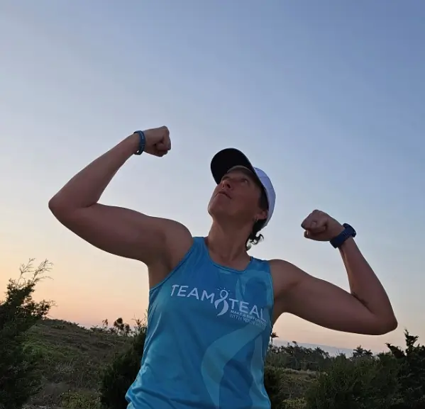 Hollie Nelson strikes a victory pose after finishing her Boston Marathon training, feeling strong and ready to race. Hollie Nelson flexes both arms in a victory pose against a sunset sky after completing her final Boston Marathon training run wearing her NOCC Team Teal shirt.